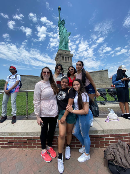 Five female high school students pose with their teacher, Ms. Dalphond, in front of the Statue of Liberty.
