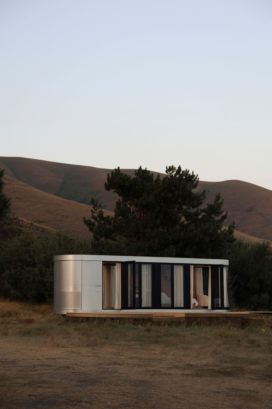 The LAVVU XL is a minimalist modular home designed by Vladislav Kulikov and manufactured by LAVVU. It features a 22 m² (237 ft²) living space built on a heavy-duty metal frame. The structure requires no concrete foundation, using hydraulic auto-leveling legs with 500 mm of adjustment to leave zero environmental footprint. It is engineered for all four seasons with energy-efficient infrared underfloor heating. Premium features include built-in home cinema projectors, automated storage, and external steel shaders to protect the large panoramic glass panels.
The brand offers four models:
- Module S: 10 m² (108 ft²)
- Module M: 14 m² (151 ft²)
- Module L: 18 m² (194 ft²)
- Module XL: 22 m² (237 ft²)
LAVVU, delivers luxury on-grid and off-grid spaces globally. Specializing in premium, fully equipped houses, LAVVU integrates contemporary architecture with intelligent engineering and well-designed interiors. Each model prioritizes functionality, quality materials, and efficient space use, ideal for private residences, guest houses, hospitality units, retreats, and commercial projects.
The kitchen is fully equipped with a built-in refrigerator, dishwasher, two-burner induction cooktop, and integrated sockets. The bathroom features a wall-hung toilet, hygienic hand shower, full-size bathtub, and clean, minimal design. The bed is positioned to maximize the panoramic view, with built-in drawers and wall shelves for personal items, along with bedside lighting and sockets.
Integrated storage throughout the LAVVU includes a wardrobe with hanging space and dedicated shoe storage, ensuring organization.
LAVVU ensures every detail, from design to custom furniture, is meticulously considered. The brand offers worldwide shipping and tailored, fully furnished setups that encourage slow living, digital detoxing, and environmental connection.
Project credits
Architects: Vladislav Kulikov @kulikovv_
Photography: Svetlana @by.sveti
Manufacturer: LAVVU