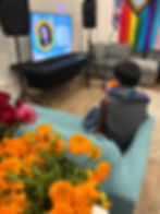 A picture of a community member holding a cup of tea and watching a slideshow of trans community members who died this past year due to violence. Marigolds and roses are in the foreground of the picture.