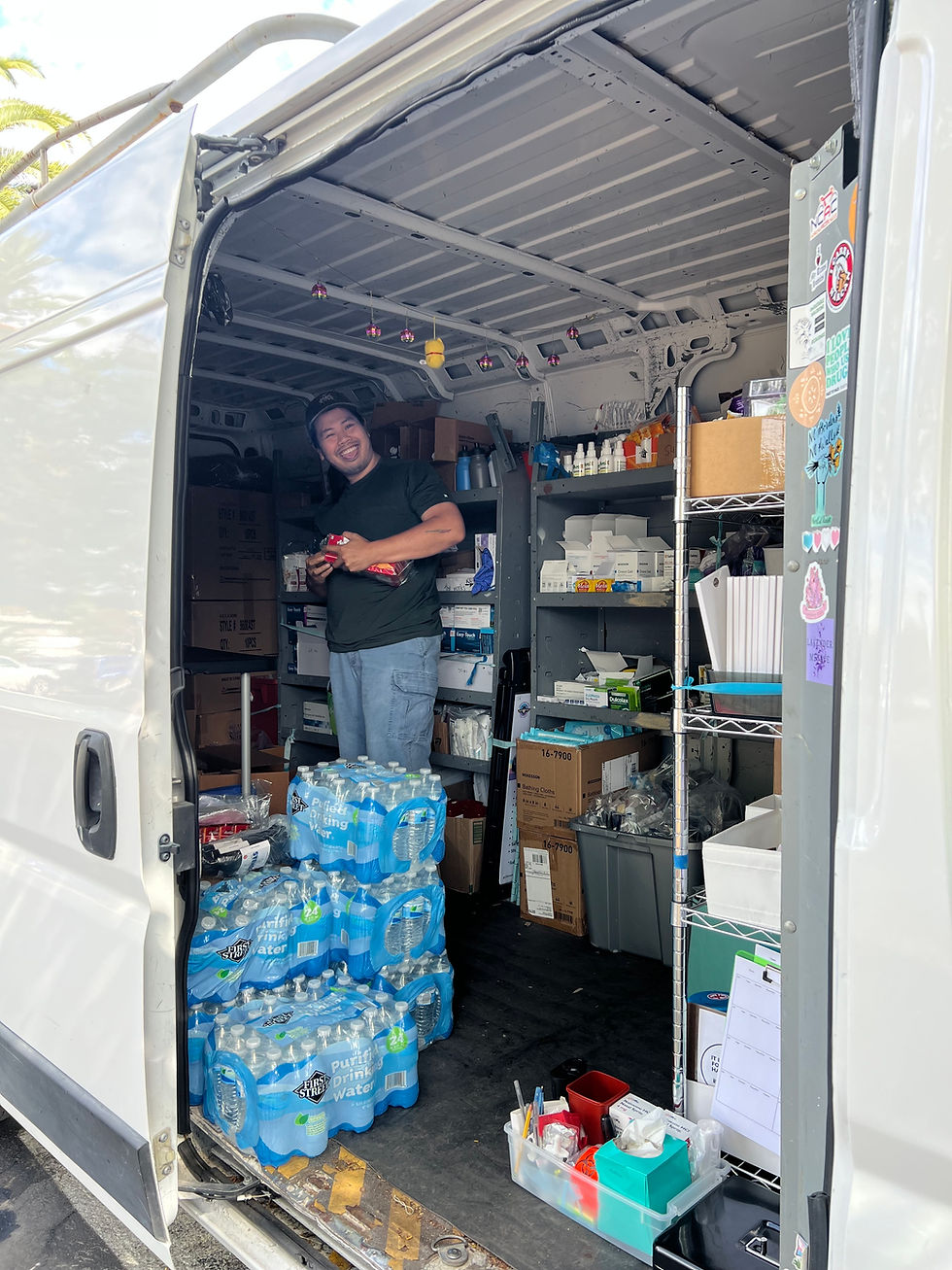 Smiling person stands in a supply-filled van, holding snacks; stacks of water bottles and sticker-covered shelves line the interior.