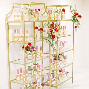 Gold geometric shelving unit adorned with vibrant floral arrangements and an assortment of pink bottled drinks, perfect for elegant event decor