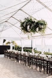 Elegant outdoor wedding setup with transparent tent, white draping, and dark wooden chairs arranged for ceremony.