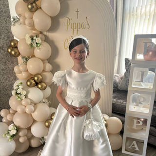 Girl smiling in a satin communion dress with lace trim and puffed sleeves, holding a satin pouch, standing indoors next to a cream and gold balloon arch and personalized "Pippa’s Communion" backdrop.