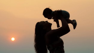 Silhouette of a woman lifting a smiling baby against a warm, orange sunset. The mood is joyful and serene.