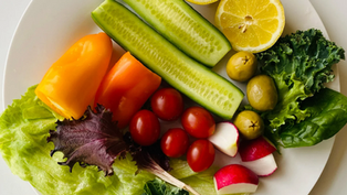 A white plate with fresh vegetables: sliced cucumber, lemon halves, cherry tomatoes, lettuce, olives, radishes, and bell peppers. Bright and colorful.