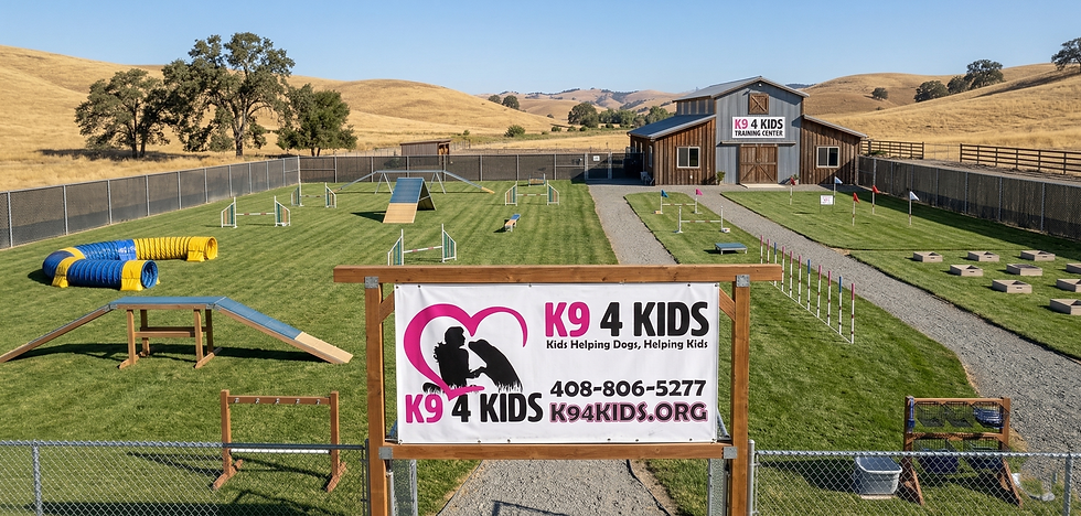 Dog training center with agility course on green grass. A sign reads K9 4 Kids, Kids Helping Dogs, Helping Kids. Hills in the background.