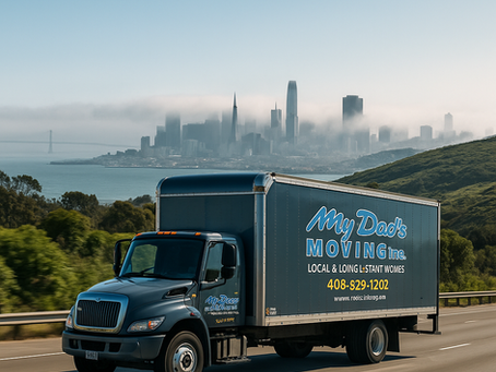 A dark gray My Dad’s Moving Inc. box truck drives on a highway flanked by green rolling hills under bright sunshine. In the distance, the San Francisco skyline rises through a low layer of fog, with the Transamerica Pyramid and Salesforce Tower faintly visible. The truck’s logo and phone number appear clearly on its side panel.