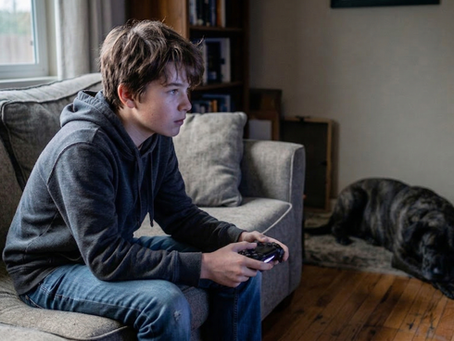 A teenager experiencing digital isolation while playing video games and ignoring a large rescue dog in the background, illustrating the necessity for screen time replacement therapy and youth canine rehabilitation programs.