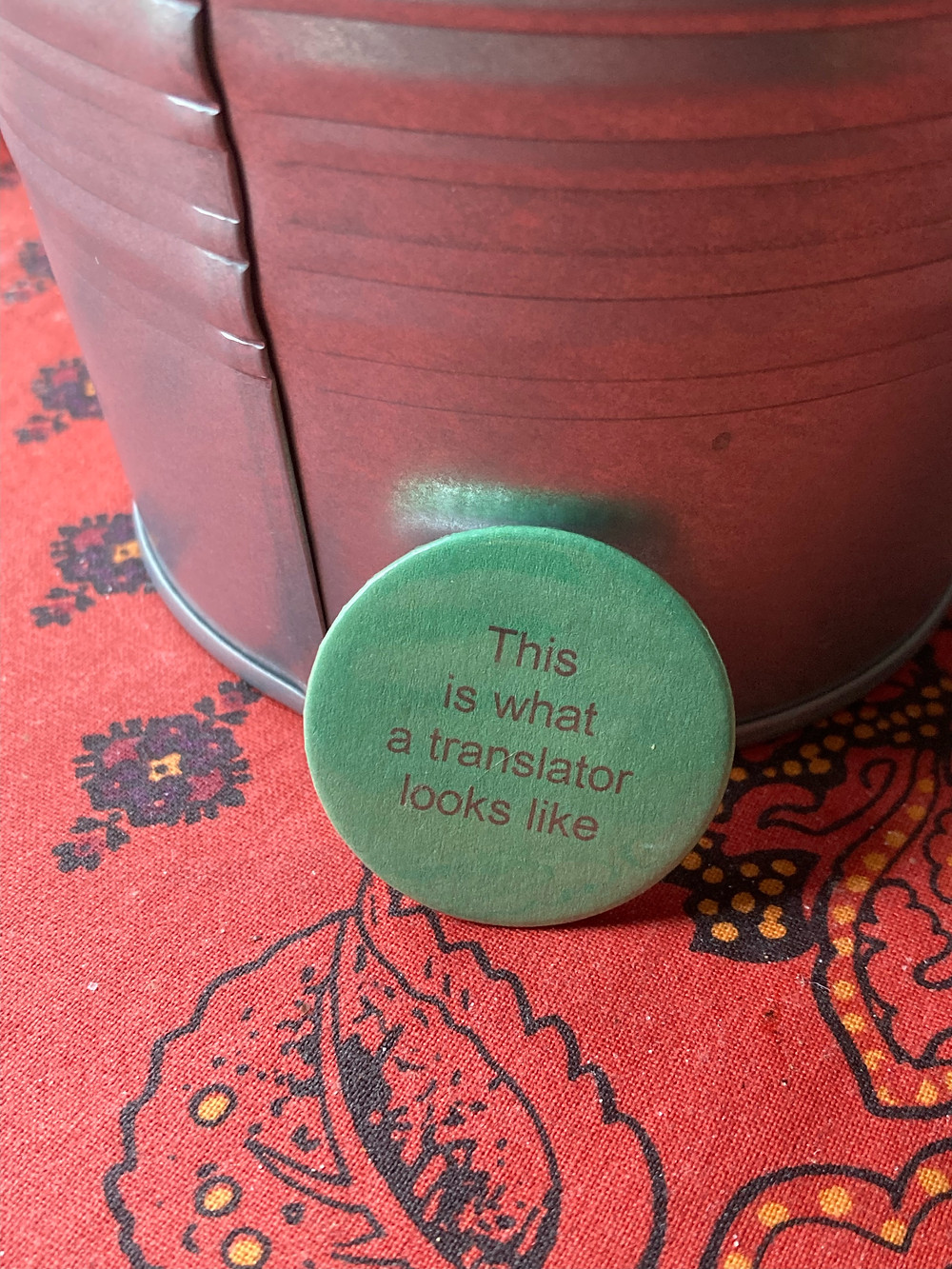 A green pin with the slogan "This is what a translator looks like" sits in front of a metal plant pot. They both sit on a red table cloth covered in a leaf pattern.