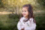 Young girl with long brown hair smiling outdoors in a white embroidered cardigan.