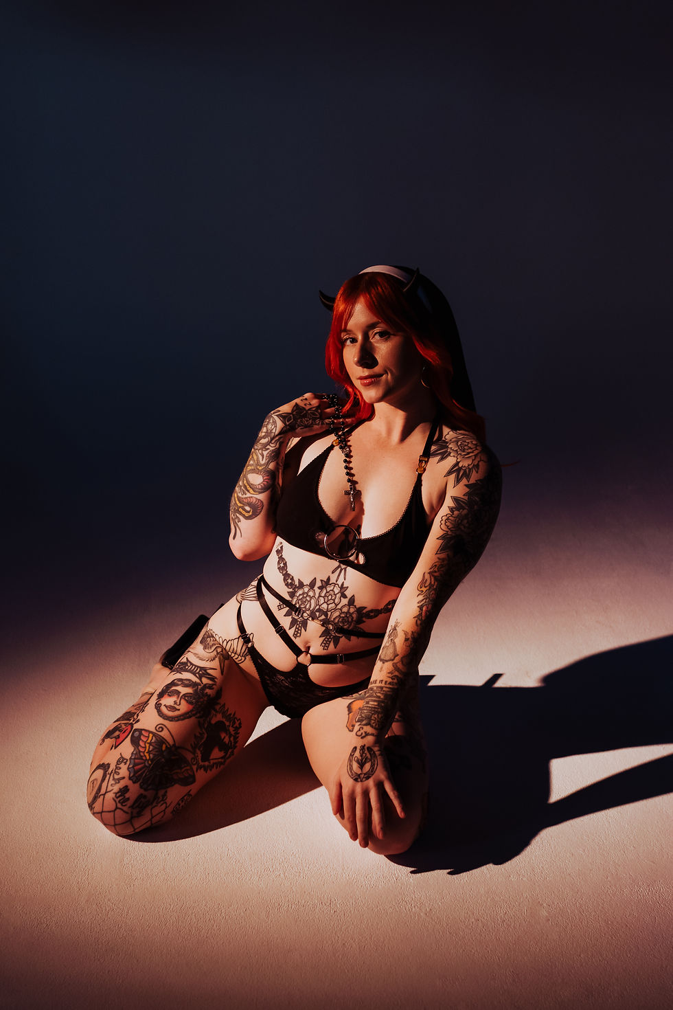 Tattooed red-haired woman in black lingerie and devil horns kneels in dramatic studio light, posing against a dark backdrop.