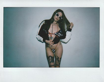Woman posing in sunglasses and a black-and-white jacket over lingerie, tattoos visible, against a plain gray background.
