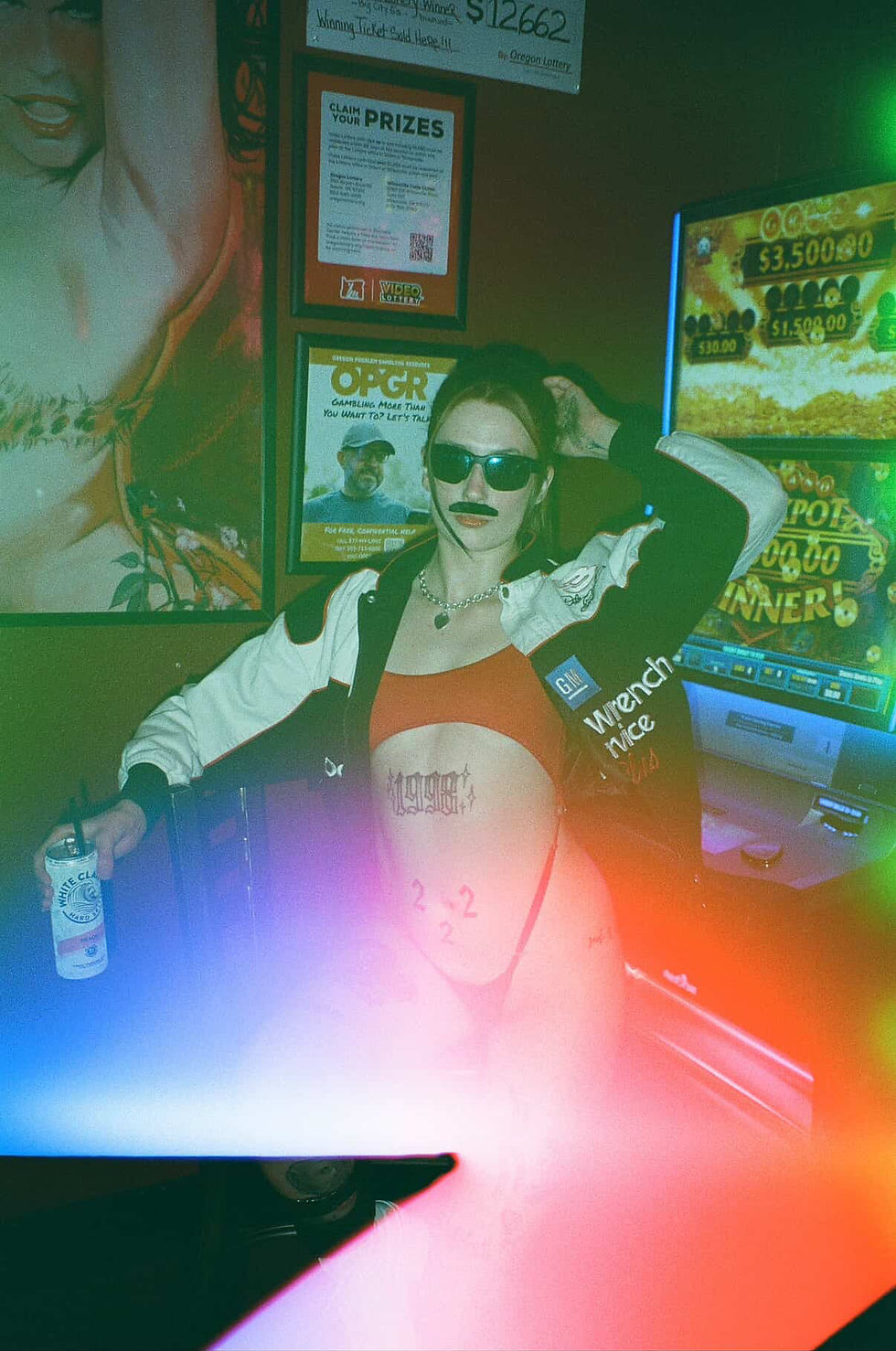 Woman with sunglasses and a fake mustache poses in a bar with colorful lights. She holds a drink, and lottery ads are visible behind her.