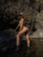 Tattooed woman in bikini and cap poses on dark rocks in a rocky stream, sunlight highlighting wet skin and rugged cliff walls.