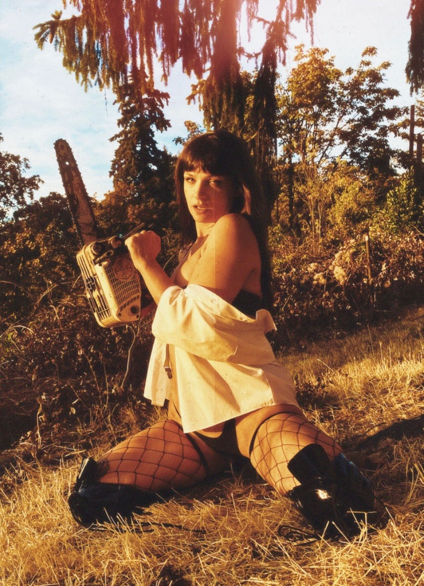 Woman kneels in dry grass at sunset, holding a chainsaw, wearing fishnet stockings, a white shirt, and black boots.