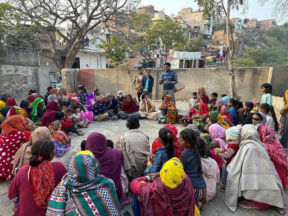 A group of women, some with their young children, sit outdoors in a circle, listening to Dr. Sharwan Saini, Founder of Save Kids Trust. Brightly colored clothes and scarves contrast with the urban backdrop.