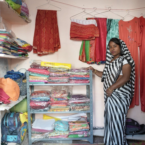 Widow showing us her business room in her house. There are many garments on shelves & some hanging