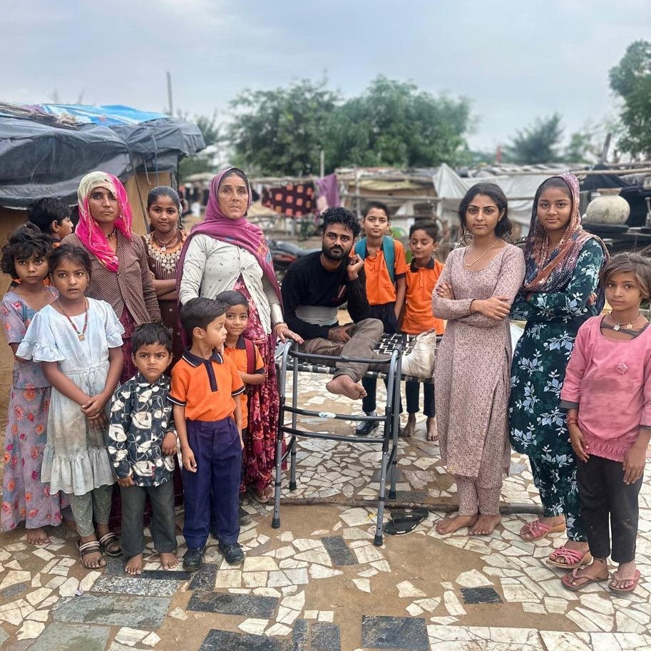 A group of people, including children in orange uniforms and adults, stand on a mosaic & dirt tiled path against a rural backdrop, appearing serious. Vimal is seated on a charpoy in the middle with his walker in front of him.