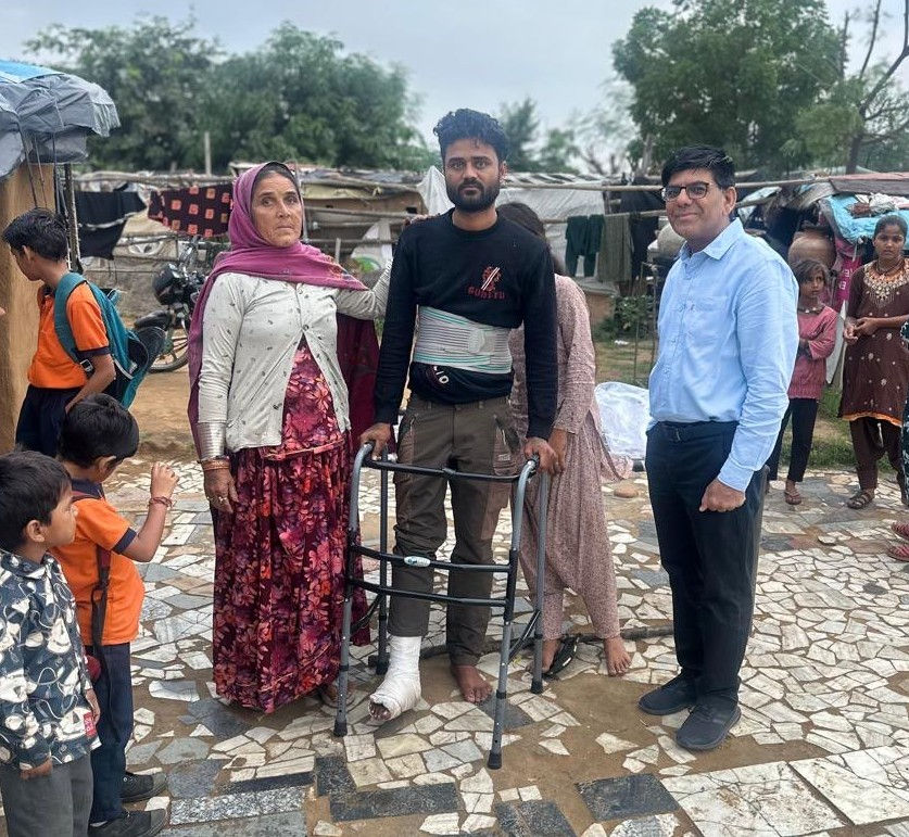 A man with a leg cast and walker stands between a woman and Dr. Sharwan Saini in an outdoor setting with mosaic tiles and children nearby.