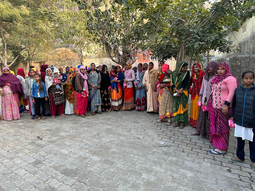 A group of women and children in colorful traditional clothing stand on a paved street under trees, holding small pink packets, looking content.
