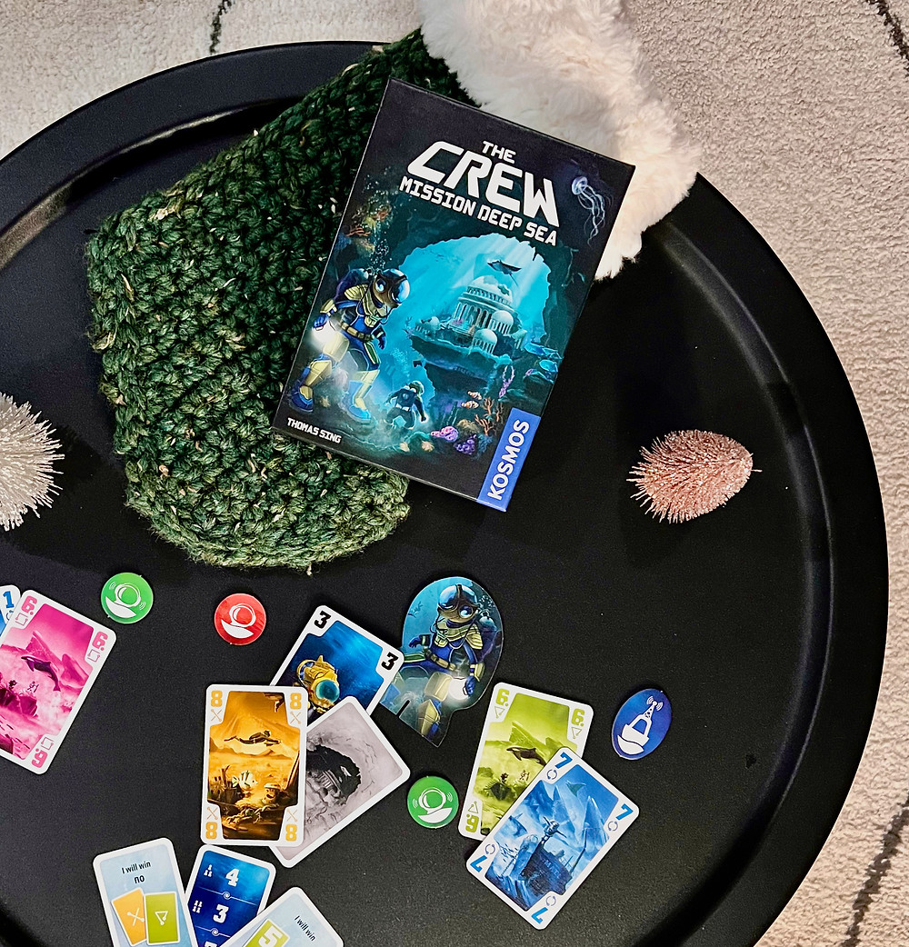 A top-down shot onto a black metal coffee table. At center, The Crew: Mission Deep Sea box is on a green crocheted stocking. A blue card with 1, and pink card with 6 are on the left. Various communication tokens are strewn throughout the frame.