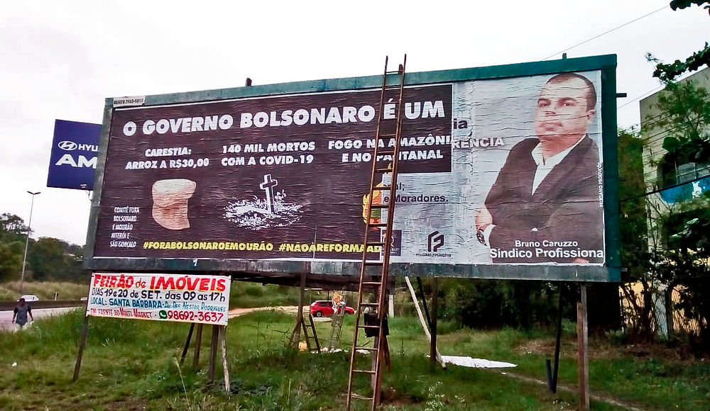 Outdoor entre rodovia e CIEP com escada escorada e imagem de uma nova mensagem aplicada pela metade. Imagem de fundo preto responsabilizando Bolsonaro pelo o aumento do preço do arroz, pelas mortes por covid e as queimadas na Amazônia e Pantanal.