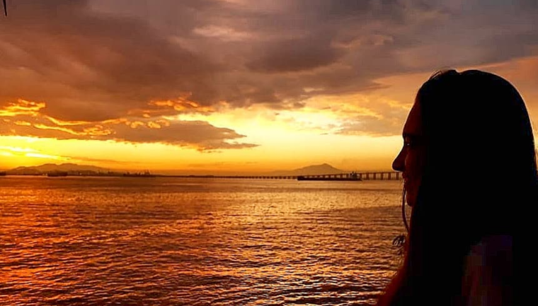 Pôr do sol com tons fortes da cor amarela e laranja em uma orla. Ao fundo a ponte Rio-Niterói e algumas embarcações. Eu estou do lado direito da imagem como uma sombra assistindo o pôr do sol.