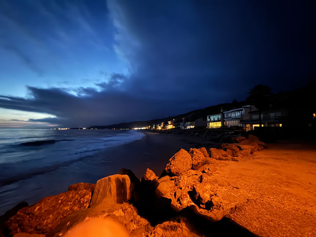 Night beach with orange rocks, waves, and distant lights