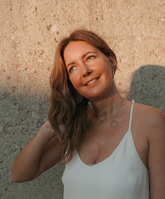 Eine brünette Frau in einem weißen Kleid lächelt in den Sonnenaufgang und streicht sich mit der rechten Hand durch die Haare