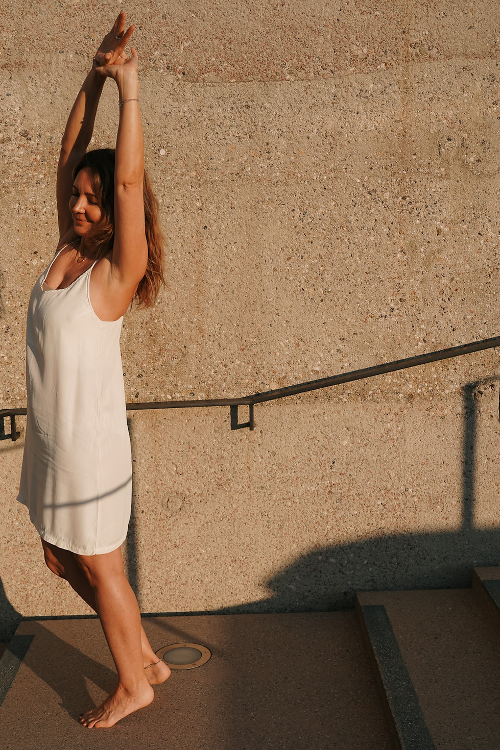 Eine Frau tanzt ein einem weißen Kleid auf einer Treppe und hebt die Arme über den Kopf während sie lächelt.