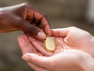 Two Black women's hands as one places a beautiful gold locket into the other's open cupped hands.