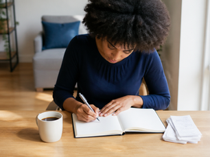 Black woman with an afro in navy sitting at a sunny table writing a list in an open journal.