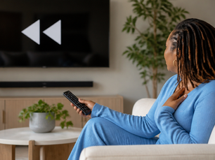 Black woman with locs in light blue top sitting on a cream sofa holding a remote pointed at a television showing a rewind symbol