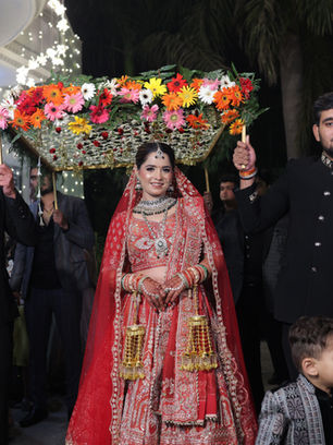 Indian bride in red lehenga walks under colorful floral canopy.