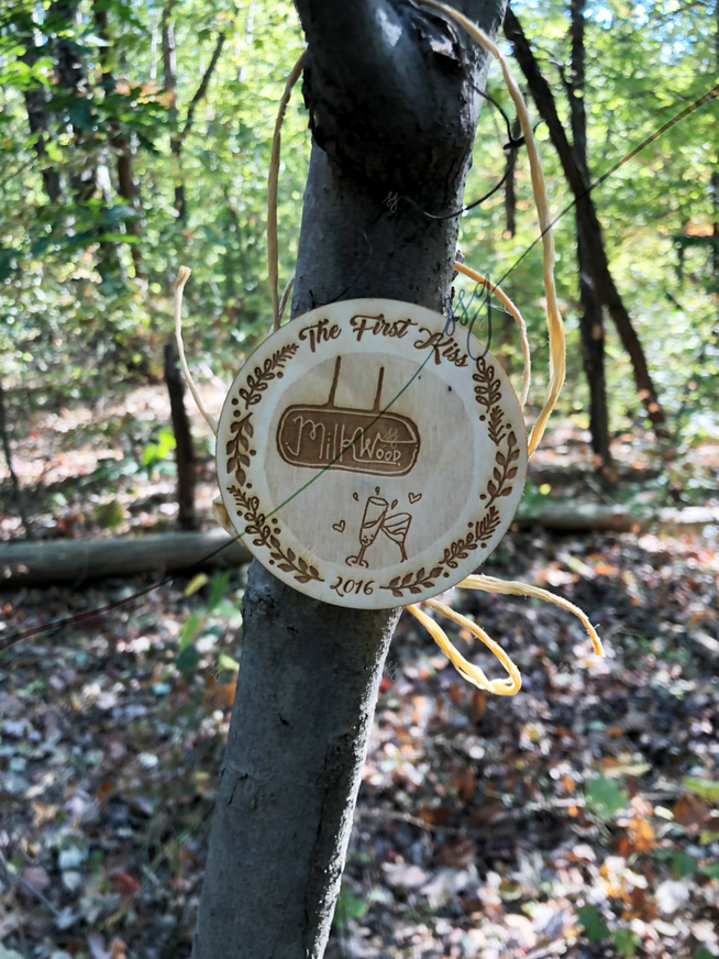 Image: A circular wooden badge laser-cut and laser-engraved with an illustration of two cocktails toasting beneath a restaurant sign and a botanical border, hanging on a tree

Text: The First Kiss