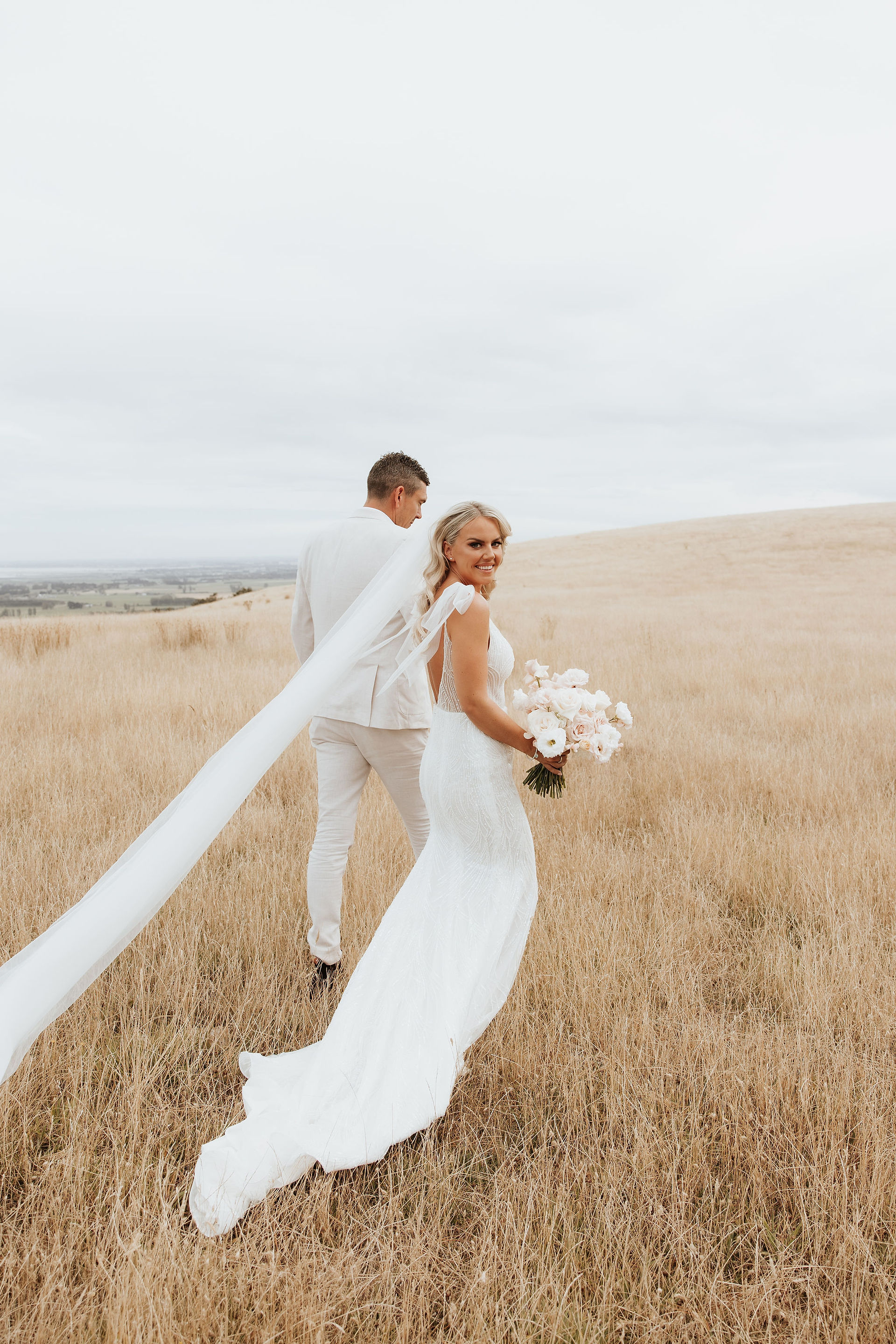 Bride's long veil trailing across the golden field as she walks beside the groom