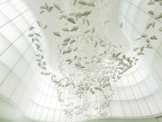 Suspended white paper-like sculptures create a delicate, floating art installation in a modern, airy space with soft light filtering through large windows.