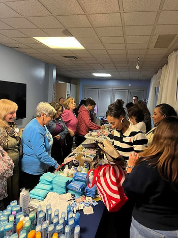 People gather around tables with hygiene products in a brightly lit room, selecting items. The mood is busy and focused.