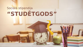 Galds uz kura stāv pulkstenis, ābols, glāze ar zīmuļiem un šķēres