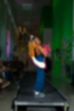 A black person in beauty pageant gear poses at the end of the runway located in a green hallway decorated with student artwork.