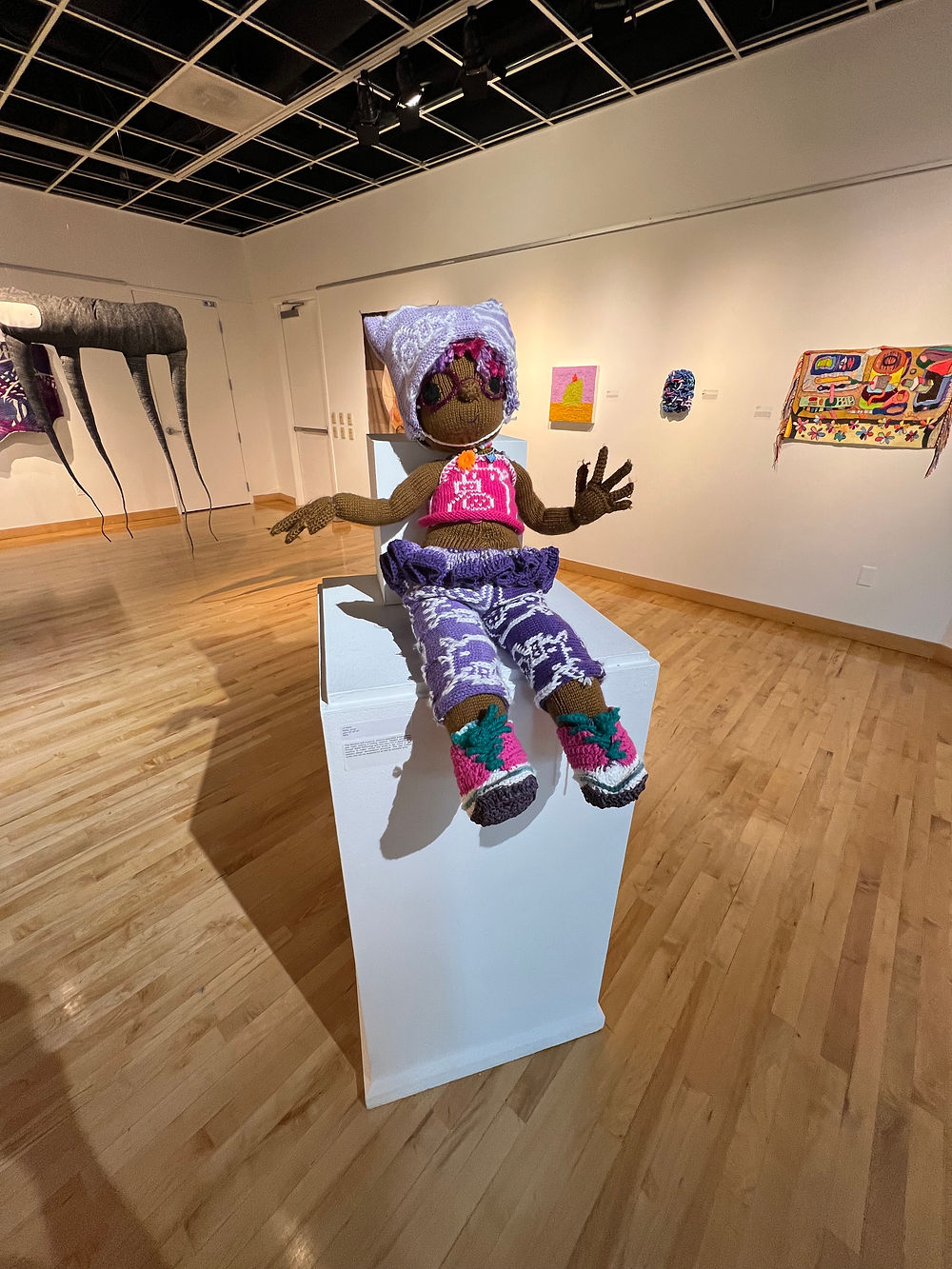 A brown-skinned knitted doll in pink and purple clothing sits posed atop a white pedestal inside a gallery with hardwood floors 