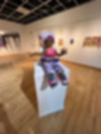 A brown-skinned knitted doll in pink and purple clothing sits posed atop a white pedestal inside a gallery with hardwood floors