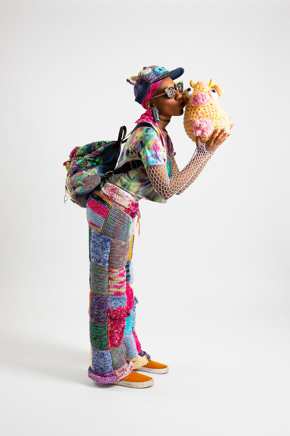 Black person wearing rainbow crocheted clothes kisses a yellow cow while standing in front of a white backdrop