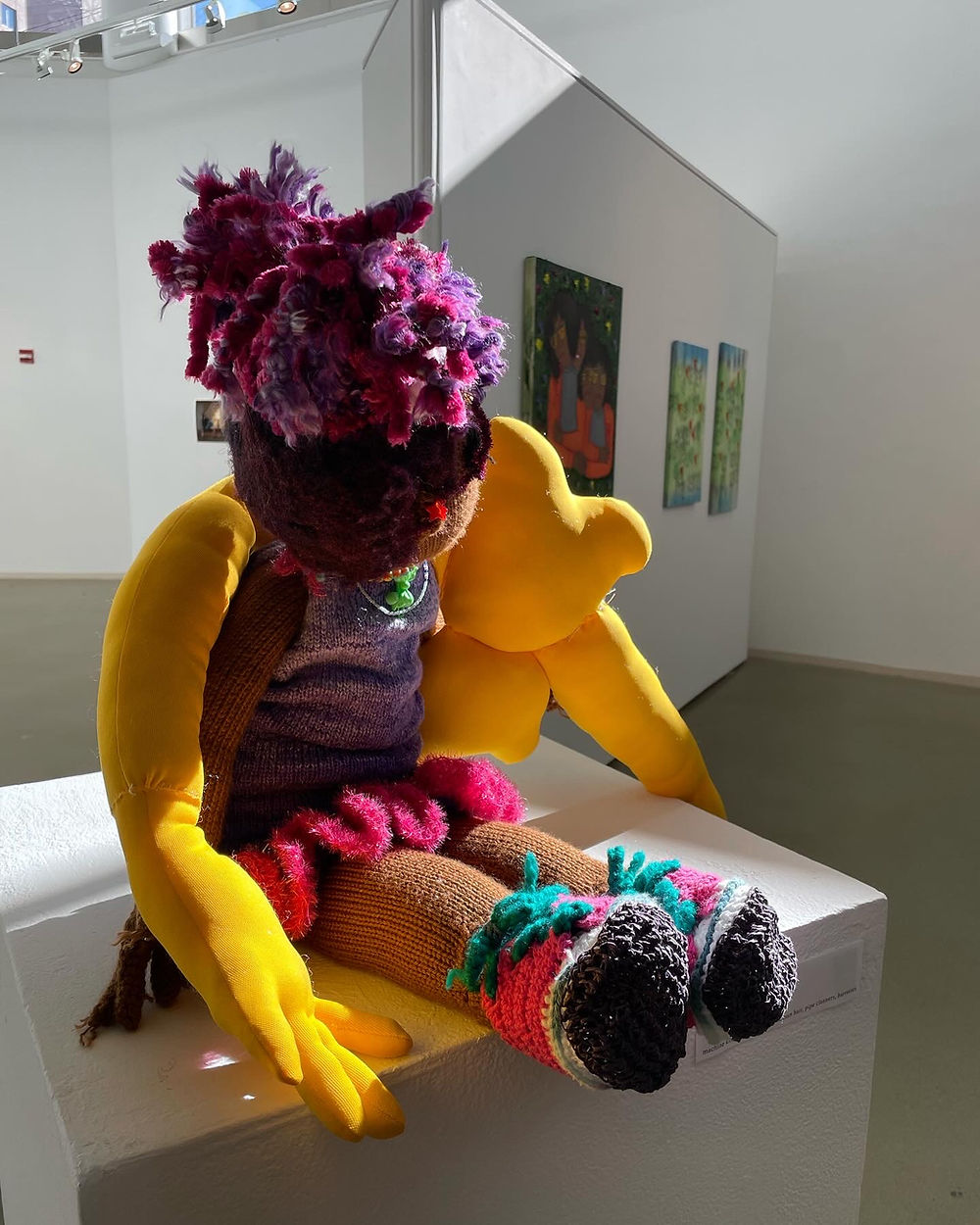 A knitted doll linking arms with a yellow stuffed form sit atop a pedestal