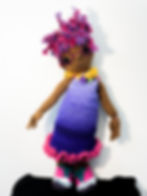 A Brown-skinned knitted doll stands in front of a white wall. She is posed to showcase her purple dress