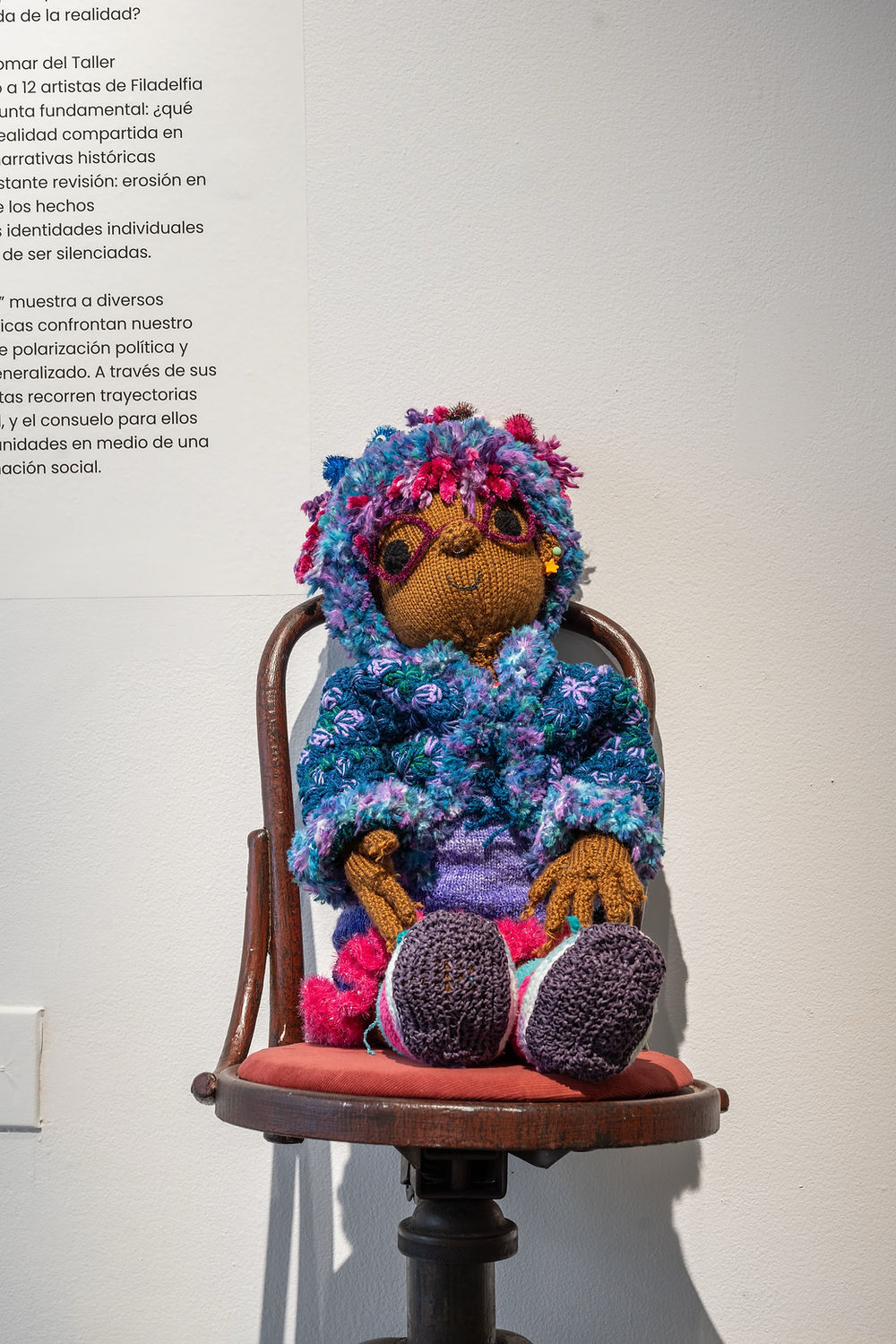 A brown-skinned knitted doll wearing crocheted clothing sits in a wooden chair positioned next to a white wall