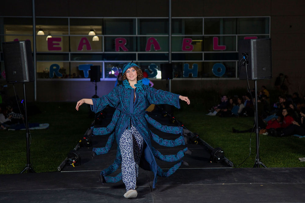 A white nonbinary person stretches their arms to so the 8 arms of a dark turquoise bug robe on an outdoor runway after dark