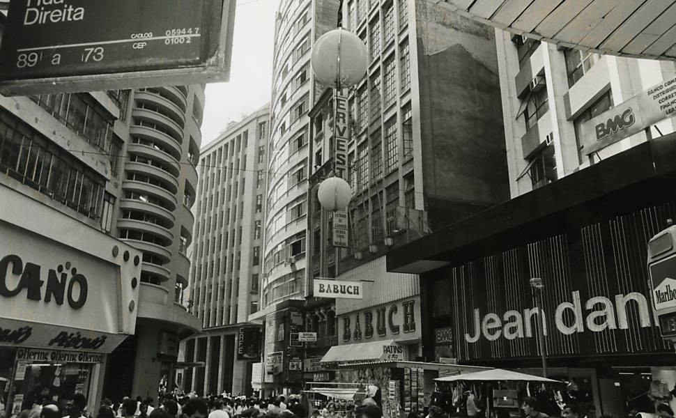 Uma rua paulista na década de 80. Uma placa escrita Direita no canto superior esquerdo (com 89 a 73). Lojas dos dois lados da rua e muitas pessoas circulando.