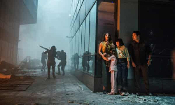 Família formada por um pai, uma mãe e duas filhas se escondendo na lateral de um edifício formado por vidros foscos pretos (no cantinho direito da imagem). No meio alienígenas armados com fuzil (em silhueta) procurando humanos com destroços de carros e outras coisas espalhadas pelo cenário. A mãe parece preocupada enquanto que a filha menor está com medo e com o rosto encostado na barriga da mãe.