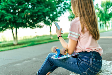 A girl sitting on a skateboard, engaged with her phone, exuding a carefree vibe in a sunny location.